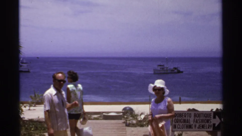 1974 Cabo San Lucas frame with a man in sunglasses, two women near the sand, offshore boats, and a Roberto Boutique sign.