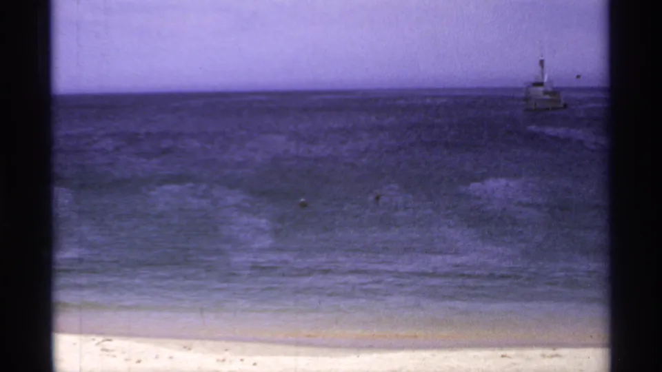 1974 Cabo San Lucas beach view showing calm blue water, pale sand, and a small boat offshore in grainy 8mm footage.