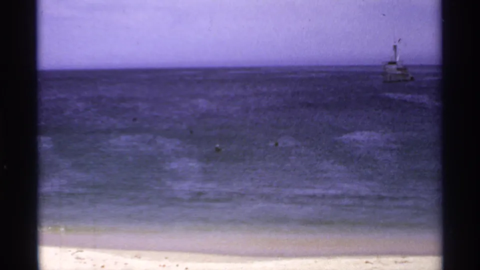 1974 Cabo San Lucas 8mm frame showing a small distant vessel offshore beyond beach, surf, and hazy sky.