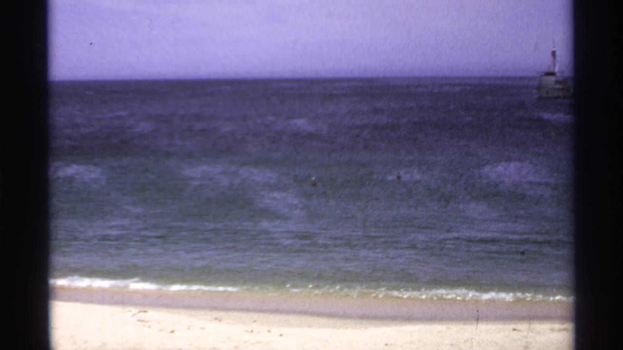 1974 Cabo San Lucas shoreline frame with a distant boat near the right edge, open water, pale beach, and faded color.