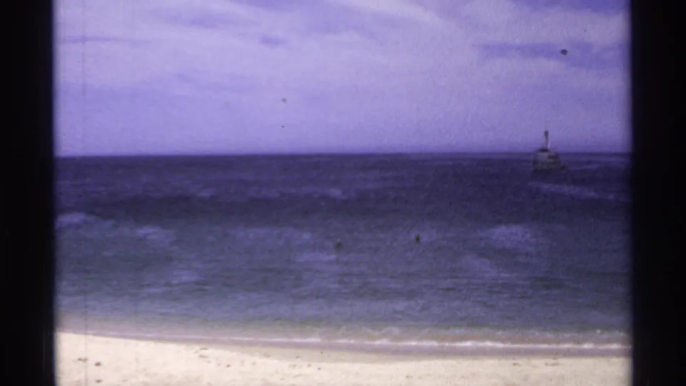 1974 Cabo San Lucas frame showing a small distant vessel offshore, with beach, surf, ocean, and hazy sky in view.
