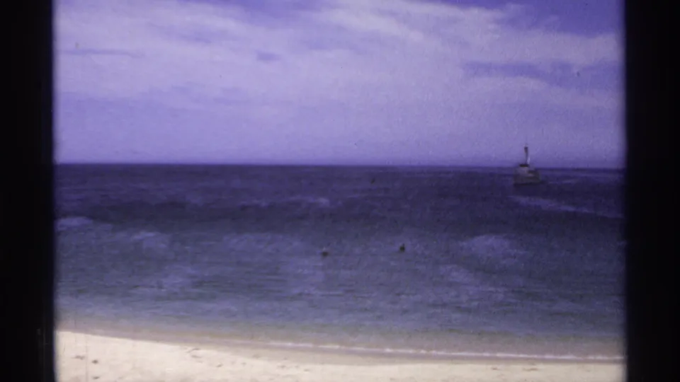 1974 Cabo San Lucas 8mm frame showing pale sand, dark water, low clouds, and a small lighthouse-like structure offshore.