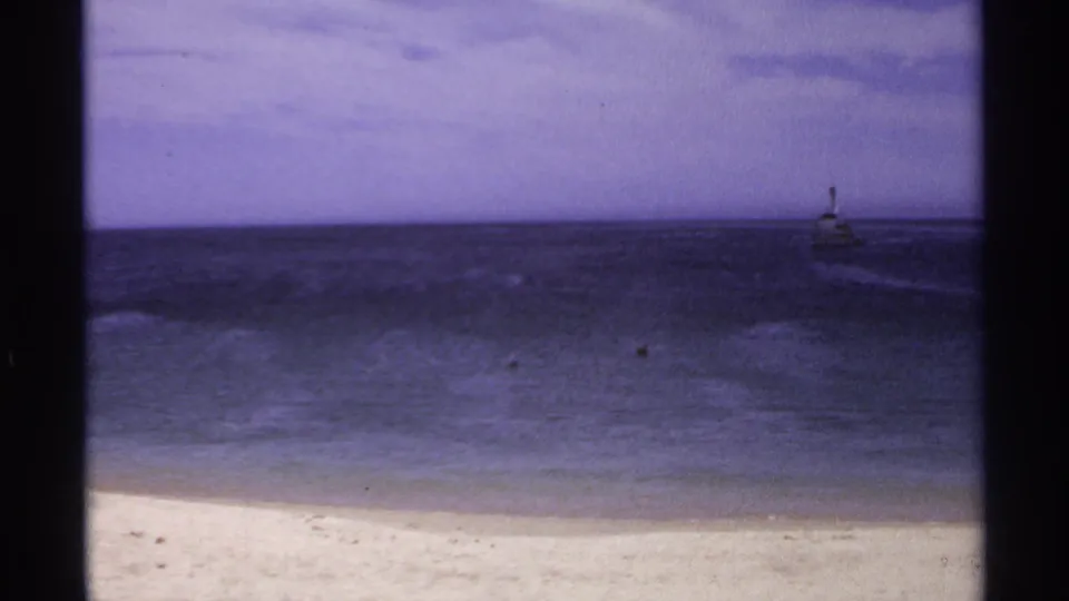 Cabo San Lucas beach view on 8mm film, with surf, dark water, purple sky, and a distant lighthouse-like form near the horizon.