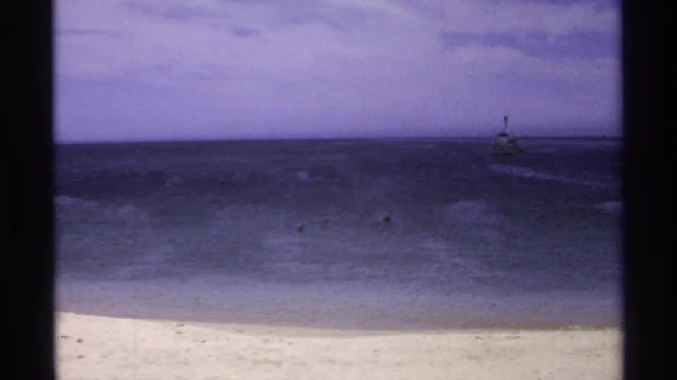 1974 Cabo San Lucas shoreline frame with pale beach, dark rippling ocean, small floating specks, and a distant offshore marker.