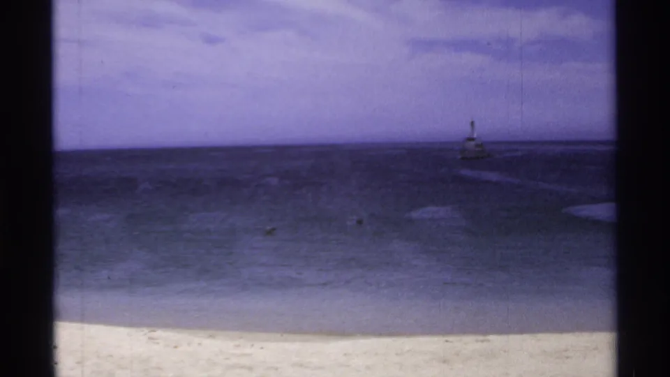 Cabo San Lucas 8mm frame showing the beach, dark open water, faint surf, and a distant lighthouse-like structure under a purple-blue sky.