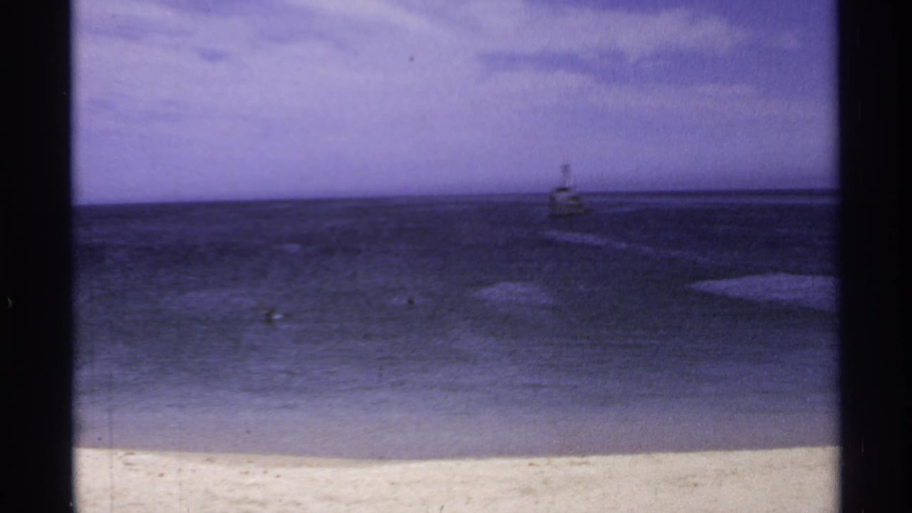 1974 Cabo San Lucas shoreline view with pale sand, dark water, clouds, and a small distant beacon offshore.