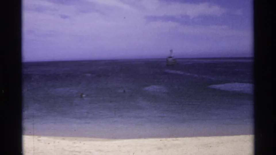 1974 Cabo San Lucas shoreline view with pale sand, dark water, clouds, and a small distant beacon offshore.