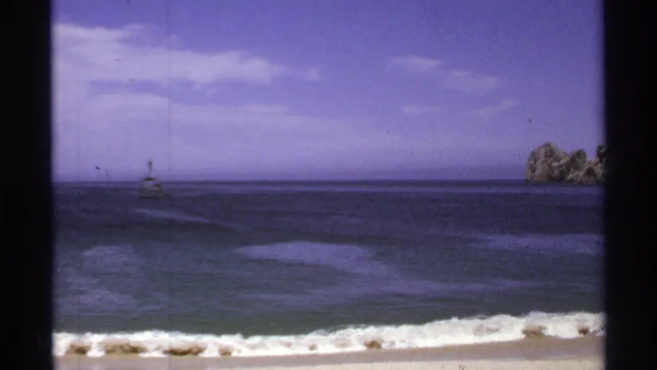Cabo San Lucas shoreline with low waves, a distant beacon, and a rocky outcrop visible on the right edge.