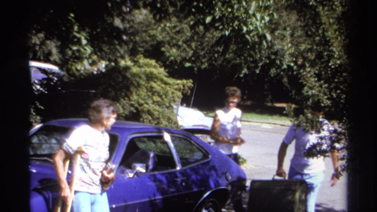 Adults stand beside a bright blue car on a sunny street with trees overhead.