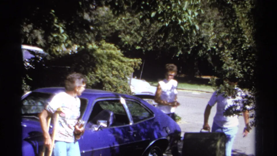 1974 La Paz home-movie frame showing adults by a bright blue car, with luggage activity beginning under leafy shade.