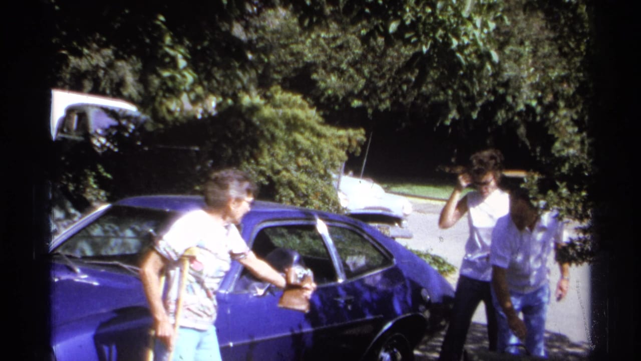 Three adults stand near the blue car as one person holds a small bag.