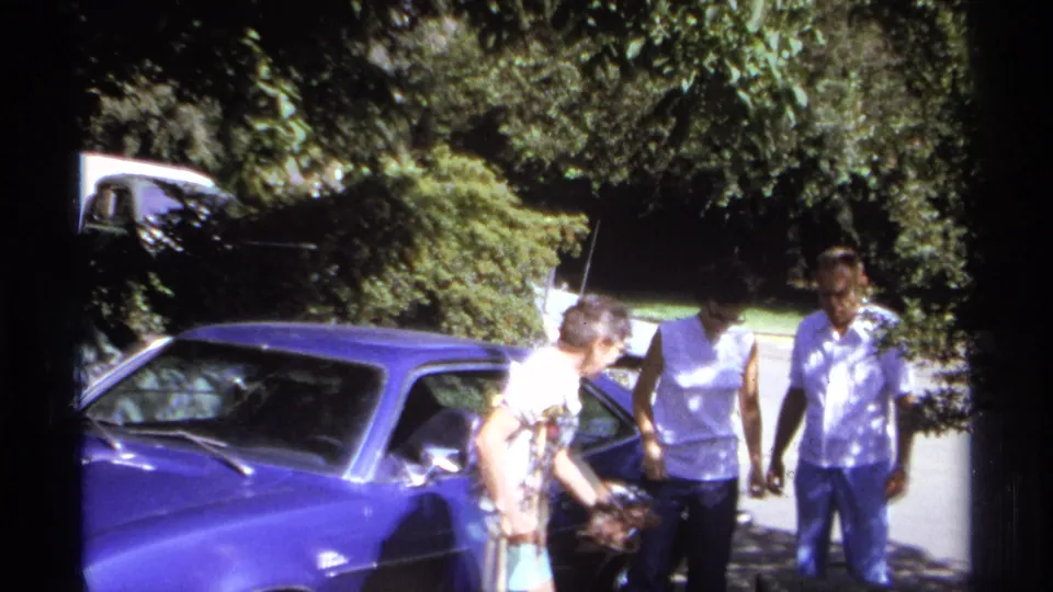 1974 La Paz frame with adults gathered by the blue car as a woman using crutches reaches toward luggage or a bag.