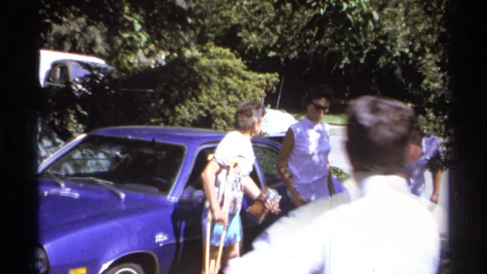 La Paz 1974 home-movie frame showing adults leaving the blue car area, with a foreground man briefly crossing the shot.