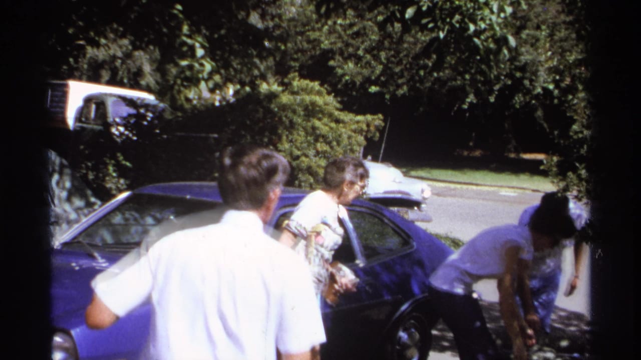 Adults stand beside a blue car on a sunny street while luggage is handled nearby.