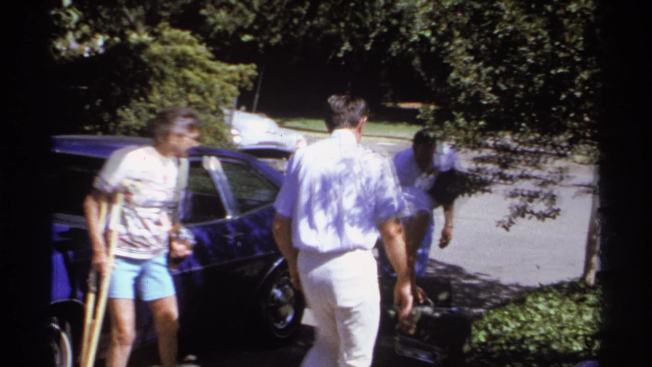 A woman with a crutch stands near a blue car as two adults move around the curbside area.