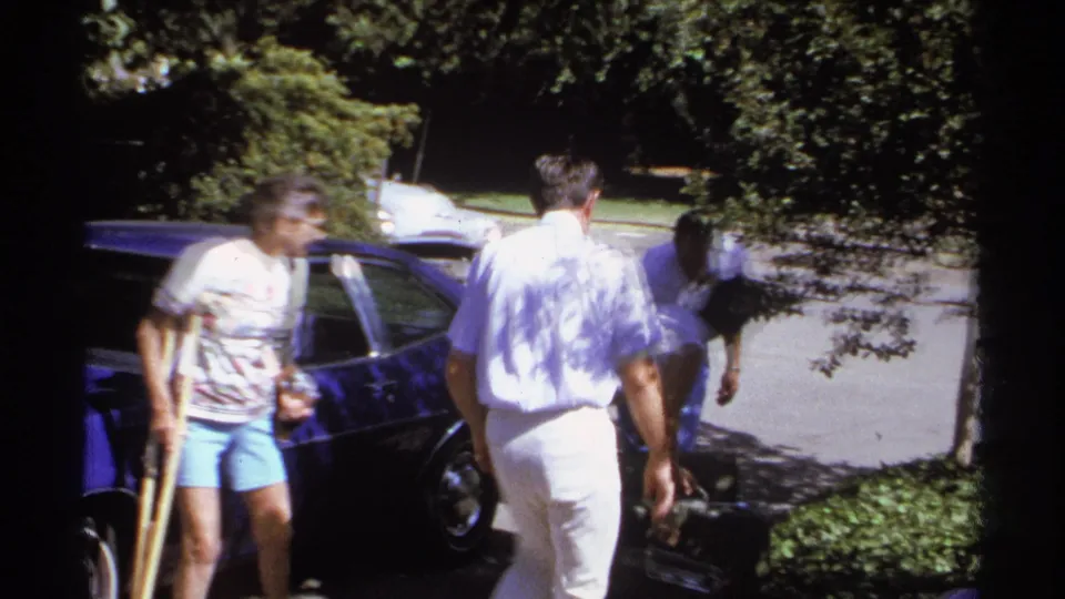 A 1974 La Paz home-movie frame shows a woman with a crutch beside a blue car while adults prepare to load belongings.