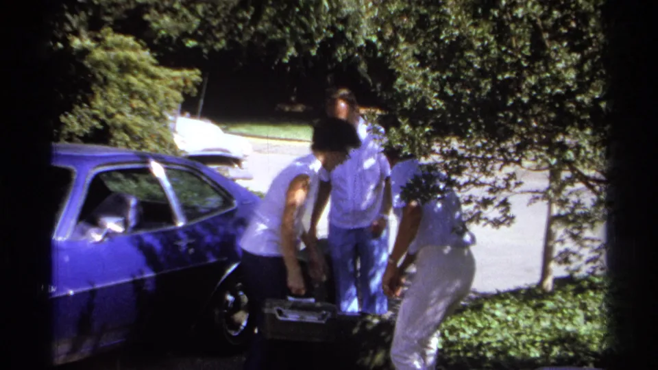 Adults in light clothing load or arrange suitcases near the rear of a blue car in 1974 La Paz home-movie footage.