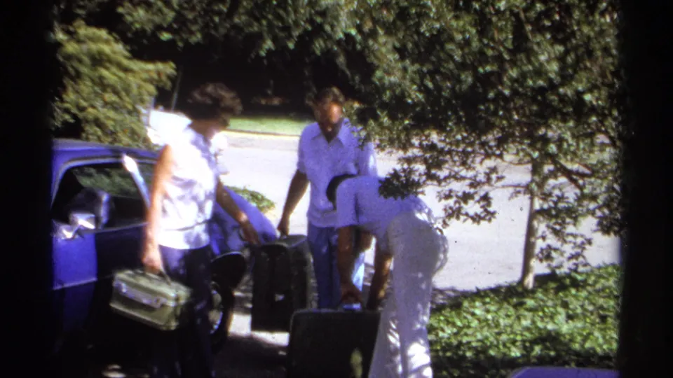 The family continues loading luggage beside the blue car in La Paz, Mexico, with a woman carrying a bag and others near suitcases.