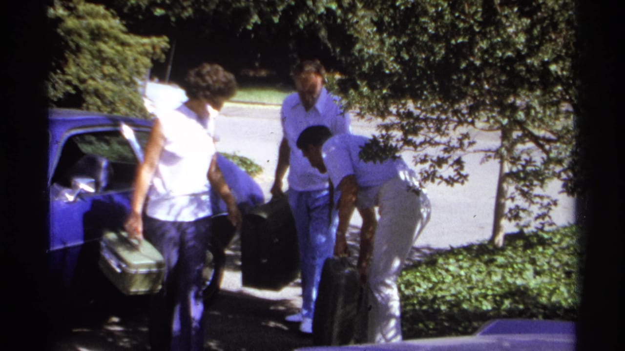 1974 La Paz home-movie frame showing adults with hard-sided luggage beside a blue sedan under street trees.