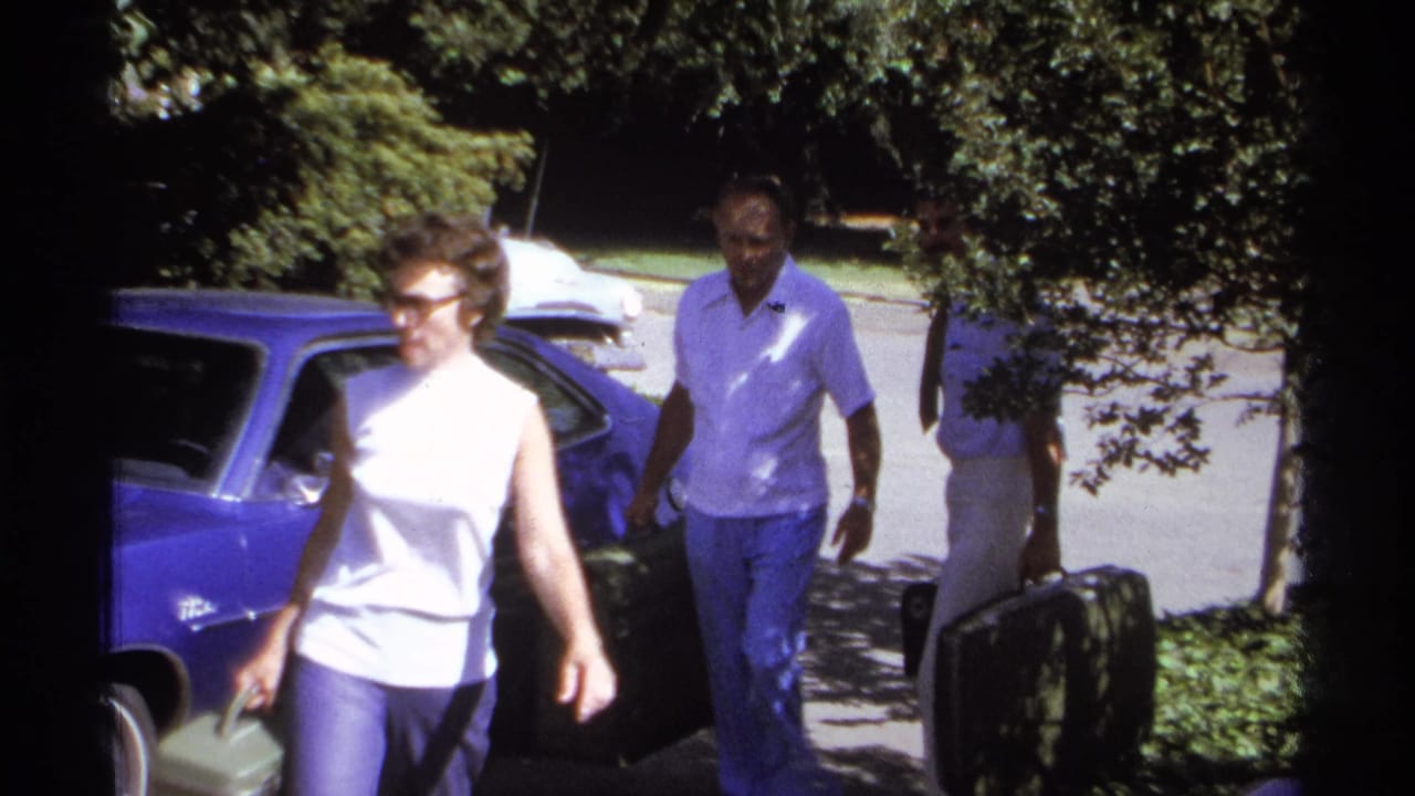Woman in sunglasses walks ahead while two men carry suitcases near a blue car.