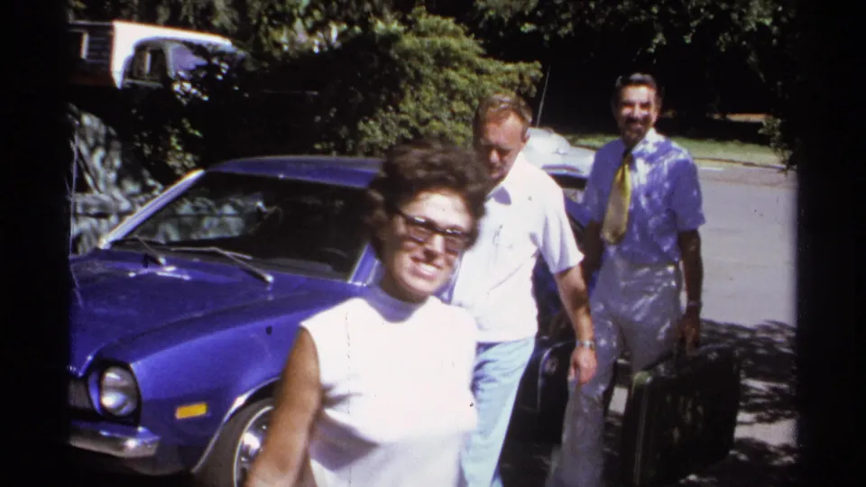 1974 La Paz home-movie frame of a smiling woman in sunglasses passing a blue sedan with men and luggage behind.