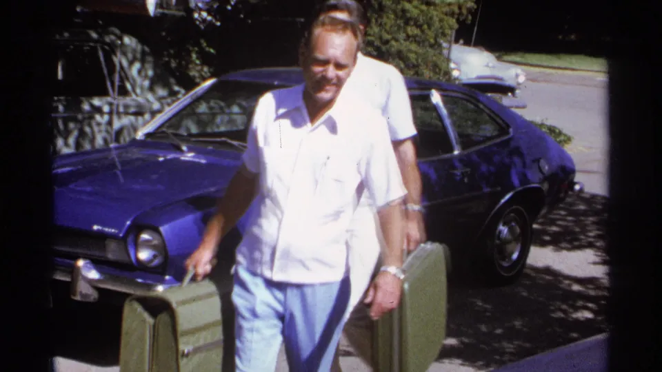 1974 La Paz frame showing an older man carrying a green hard-sided suitcase in front of a blue sedan.
