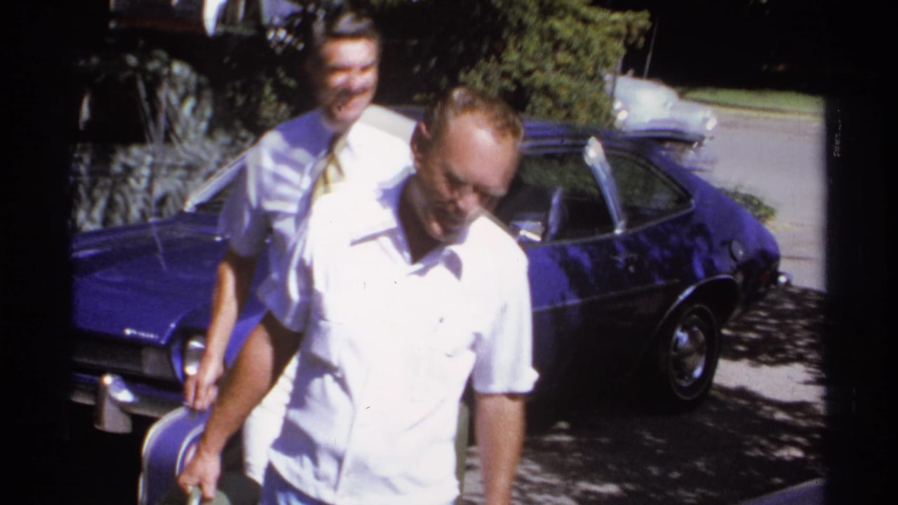 Two men walk past a blue parked car, one carrying a green suitcase.