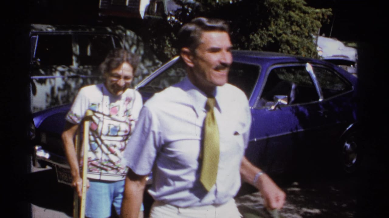 A man in a yellow tie walks ahead of an older woman using crutches near a blue car.