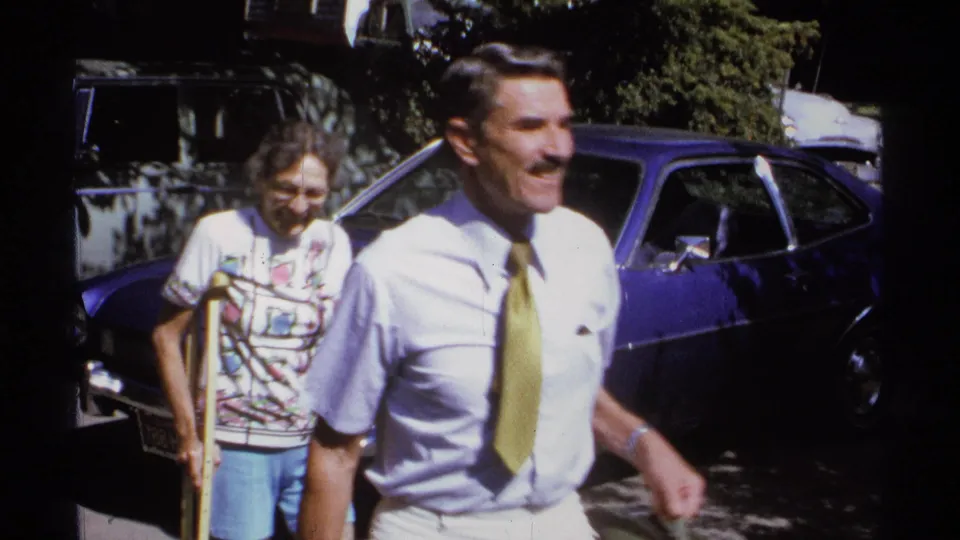 In the 1974 La Paz clip, a man in a yellow tie passes the camera while an older woman with glasses uses crutches behind him near the blue car.