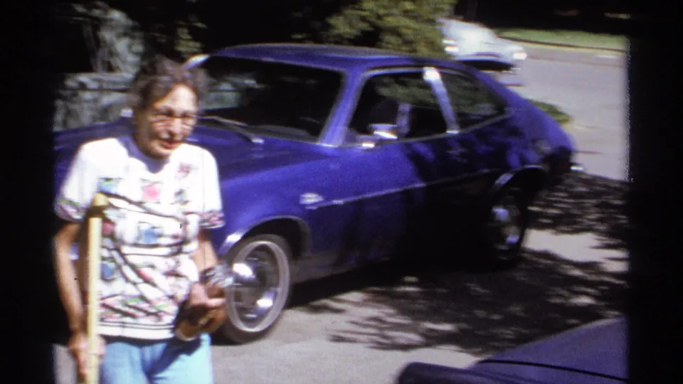 The older woman, wearing glasses and a patterned shirt, uses crutches while moving in front of a blue parked car in the La Paz street scene.