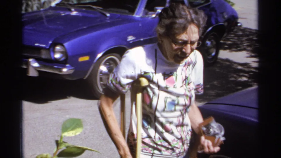 A closer frame shows the older woman with glasses and crutches passing beside the blue car, with strong sun and 8mm softness visible.