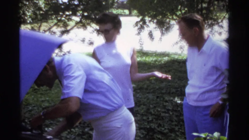 A man leans into the open trunk of a blue car while two adults in white clothing wait nearby under tree shade.