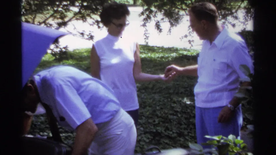 Two adults appear to talk beside the open trunk while another person bends near the car during the 1974 La Paz travel scene.