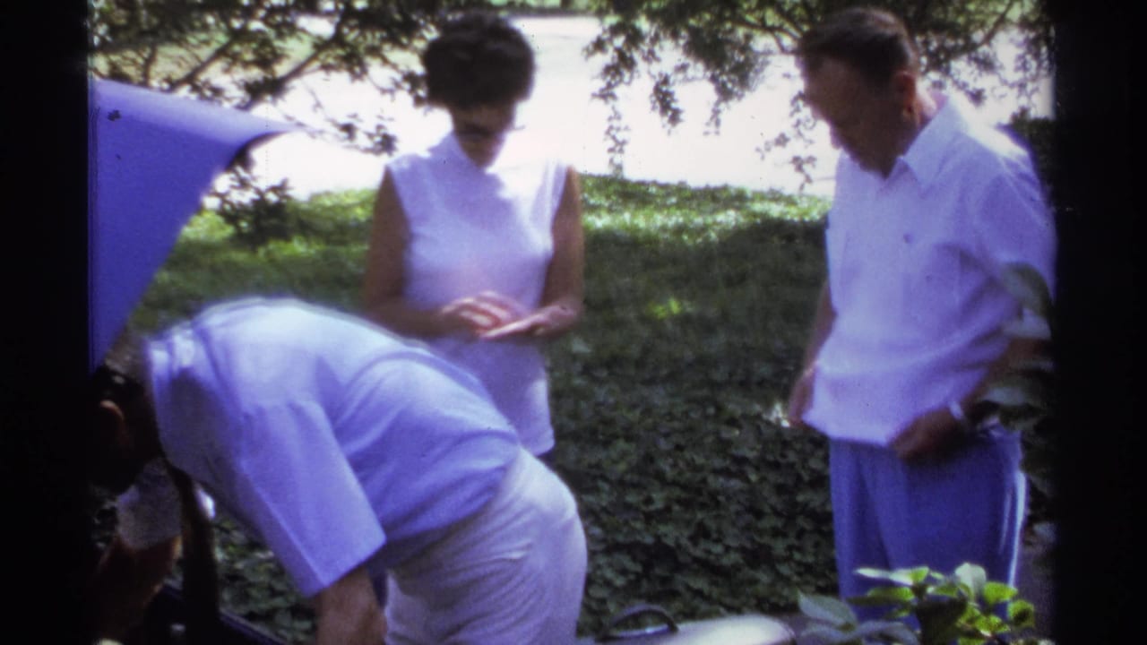 Adults in a shaded driveway stand near luggage and greenery.