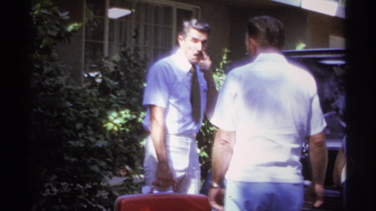 Man with cigarette stands near another man beside an open car trunk.