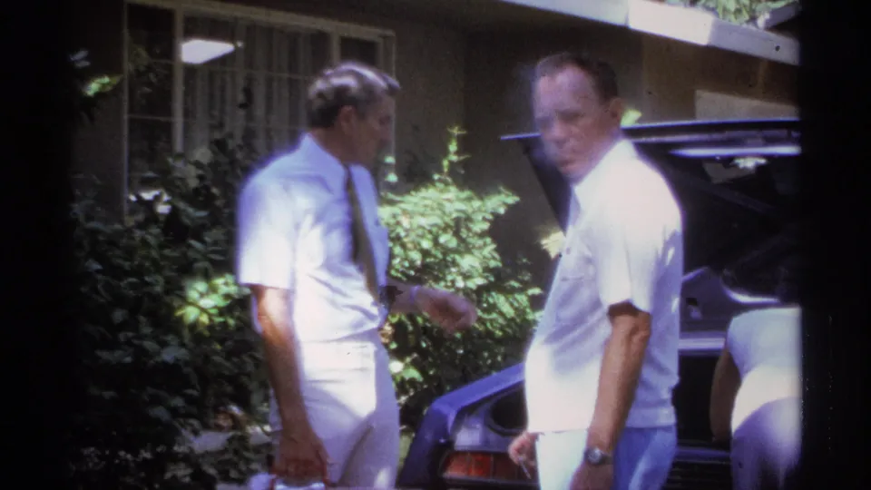 Two adult men in light shirts stand beside a car with its trunk open during a 1974 La Paz home-movie moment.