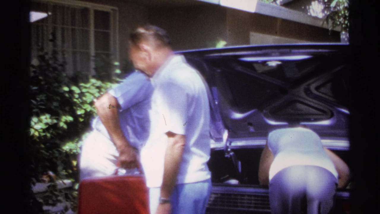 In 1974 La Paz, Mexico home-movie footage, adults in light clothing pack luggage into an open car trunk outside a residence.