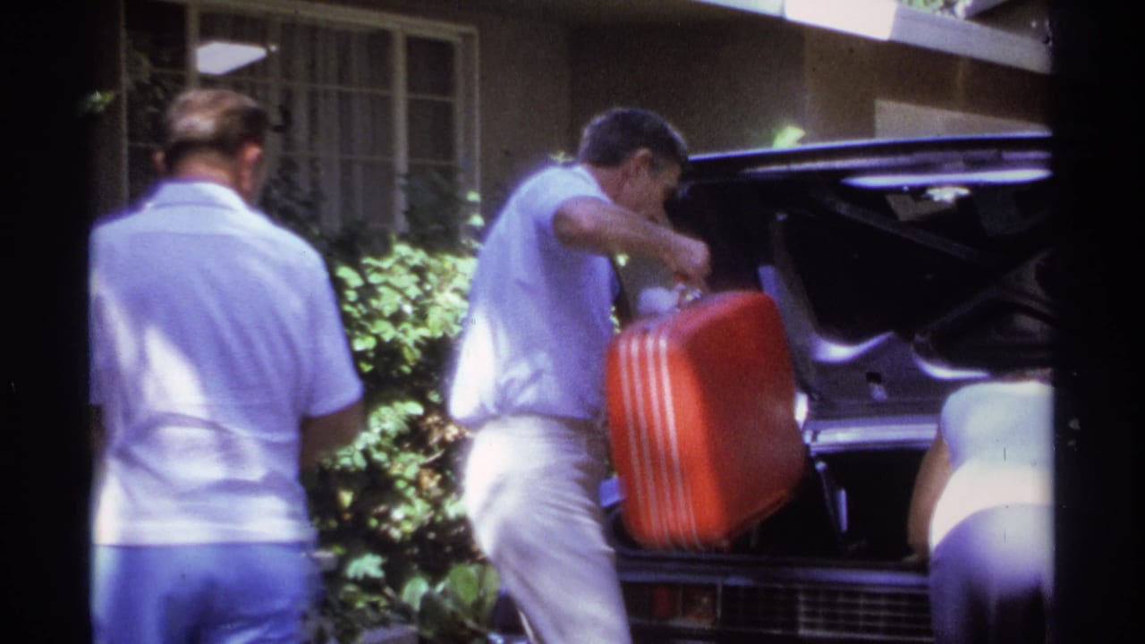 A man lifts a red suitcase toward the open trunk as another adult stands nearby.