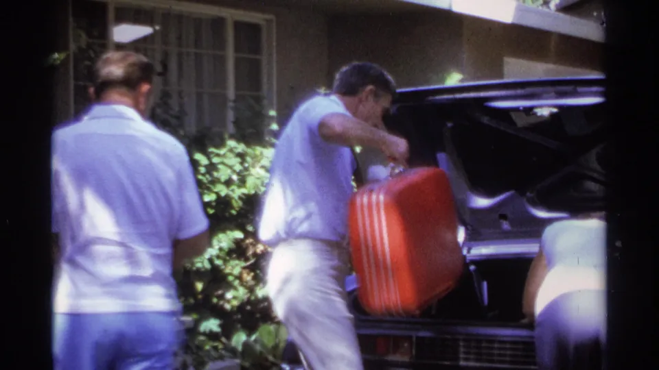 A man places a red hard-sided suitcase into a vintage car trunk while another adult stands beside him in bright daylight.