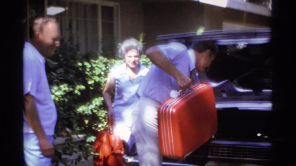 Adults in summer clothing continue packing beside the open car trunk, with a woman visible near shrubs in the residential setting.