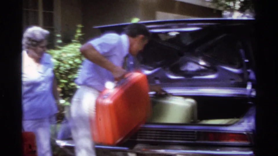 A red suitcase is lowered into the open trunk, where pale luggage is already packed, as another adult walks past.
