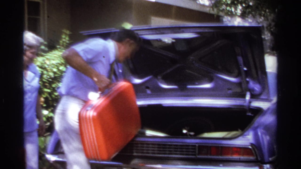 Man lifting a red suitcase beside an open purple car trunk as a woman stands nearby.