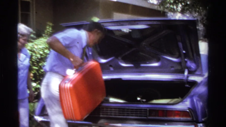 In La Paz, Mexico, 1974, a man in a light shirt lifts a red suitcase toward the open trunk of a purple car while a woman watches nearby.