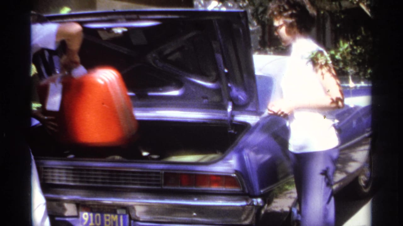 A red suitcase is lowered into the open trunk of a purple car during a 1974 La Paz home-movie travel packing scene.