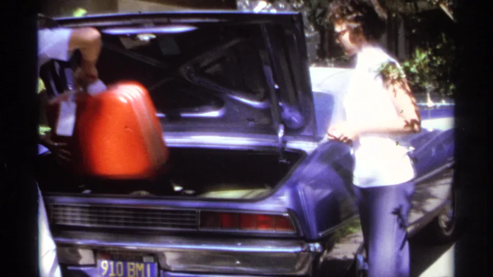A red suitcase is lowered into the open trunk of a purple car during a 1974 La Paz home-movie travel packing scene.