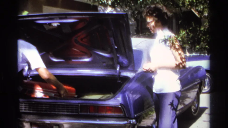An adult reaches into the open trunk of a purple car as a red suitcase sits inside during the La Paz, Mexico travel preparation clip.