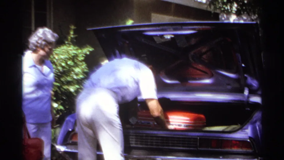 A man bends into the open trunk of a purple car to arrange a red suitcase, with a woman standing nearby in the 1974 La Paz home movie.