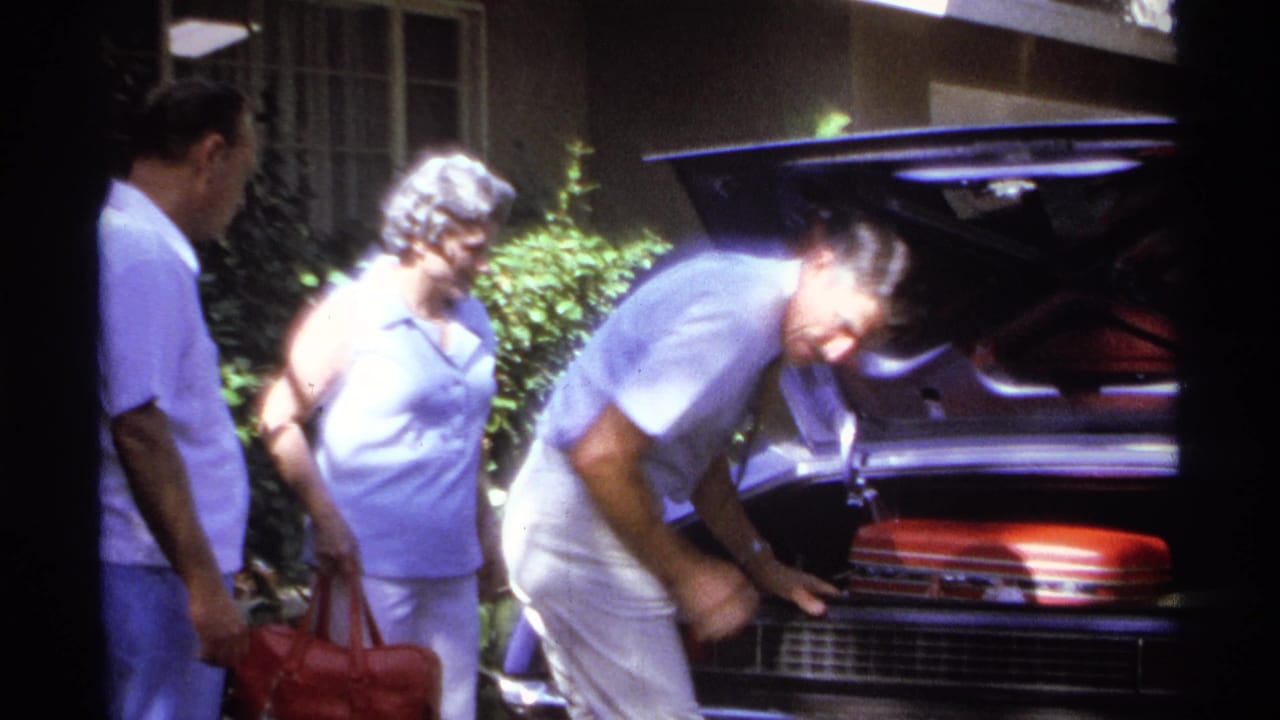 In 1974 La Paz, Mexico, adults in light clothing gather in a driveway while a man loads a red suitcase into the open trunk of a parked car.