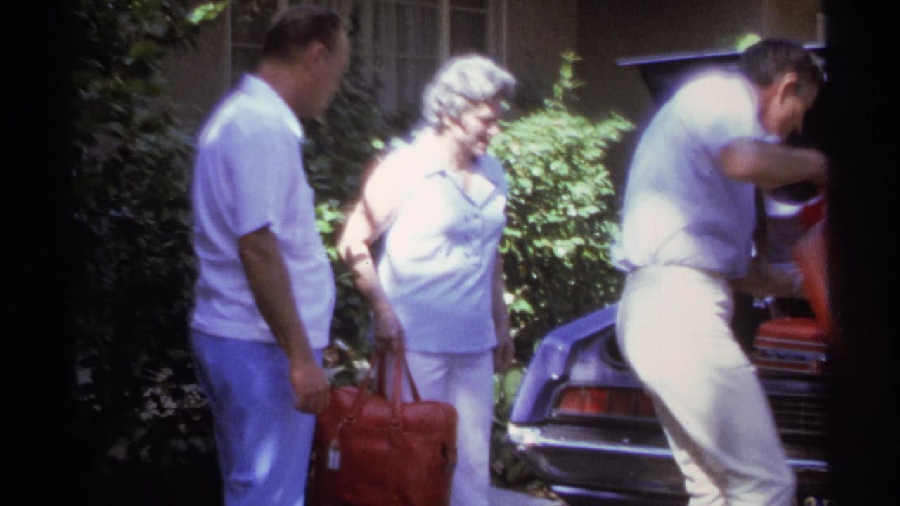 A man leans into the trunk of a classic car as another man and a woman stand nearby with a red bag in a sunlit La Paz driveway.