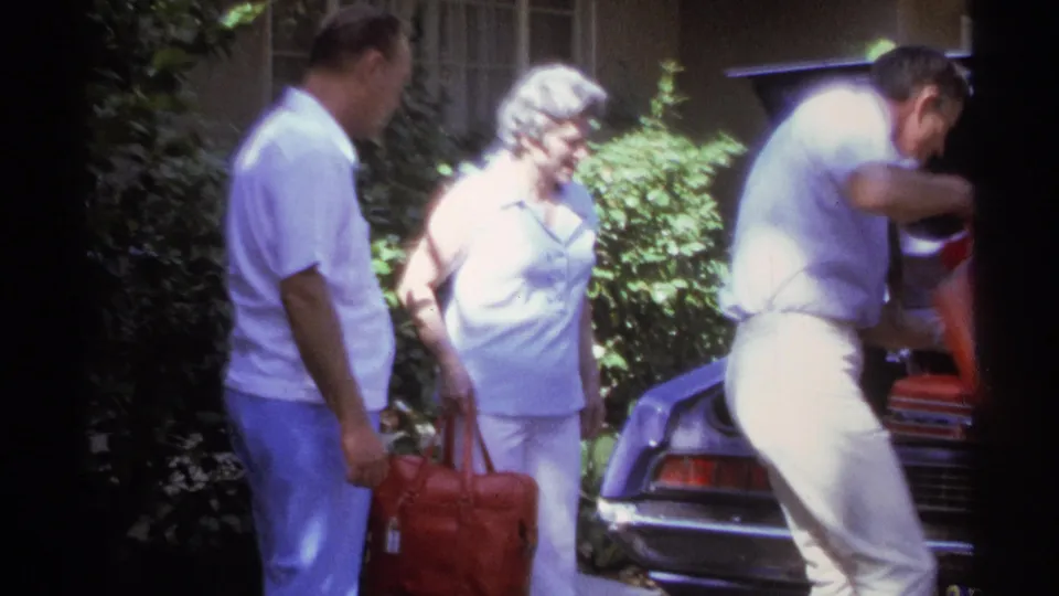 A man leans into the trunk of a classic car as another man and a woman stand nearby with a red bag in a sunlit La Paz driveway.